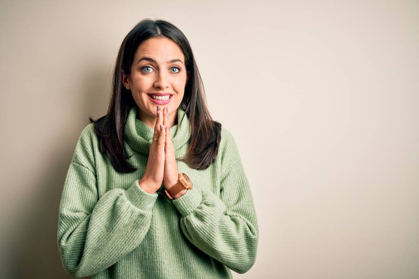 Νεαρή μελαχρινή γυναίκα με μπλε μάτια φορώντας ζιβάγκο πουλόβερ πάνω από λευκό φόντο προσεύχεται με τα χέρια μαζί ζητώντας συγχώρεση χαμογελώντας αυτοπεποίθηση. - Φωτογραφία, εικόνα