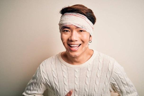 Junger gutaussehender chinesischer Mann verletzt sich bei Unfall mit Verband und Streifen am Kopf lächelnd und laut lachend, weil lustiger verrückter Witz mit Händen am Körper. - Foto, Bild
