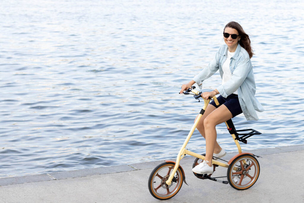 White beautiful young happy free woman rides a bicycle along the concrete promenade in the afternoon. Blue water and clothes, dark hair. She is looking at the camera. - 写真・画像