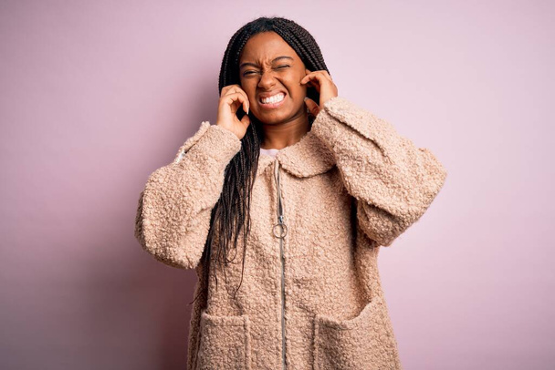 Giovane donna afroamericana che indossa cappotto invernale di moda su sfondo rosa isolato che copre le orecchie con le dita con espressione infastidita per il rumore della musica ad alto volume. Concetto di sordo
. - Foto, immagini