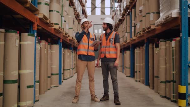 Dwóch młodych pracowników stojących w magazynie dystrybucyjnym w białym twardym kapeluszu i kurtce odblaskowej badających zapasy za pomocą tabletu cyfrowego - Materiał filmowy, wideo