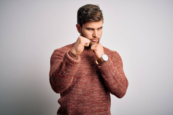 Young blond man with beard and blue eyes wearing casual sweater over white background Ready to fight with fist defense gesture, angry and upset face, afraid of problem - Foto, afbeelding
