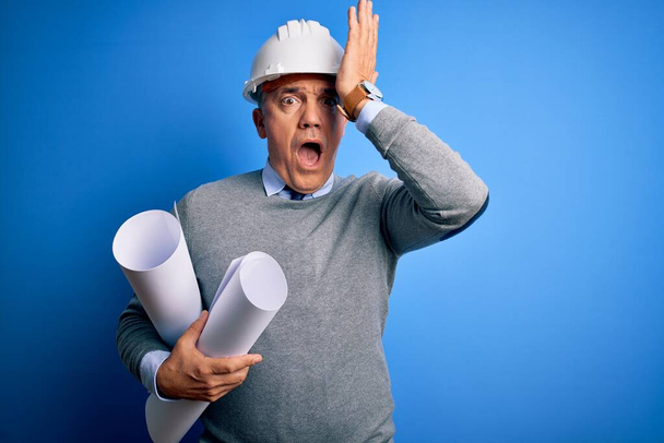 Ein gutaussehender grauhaariger Architekt mittleren Alters, der einen Schutzhelm trägt und Baupläne hält, die mit der Hand auf dem Kopf für einen Fehler überraschen, erinnere dich an einen Fehler. Vergessenes, schlechtes Gedächtnis-Konzept. - Foto, Bild