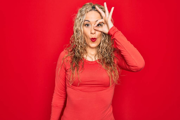 Young beautiful blonde woman wearing red casual t-shirt standing over isolated background doing ok gesture shocked with surprised face, eye looking through fingers. Unbelieving expression. - 写真・画像