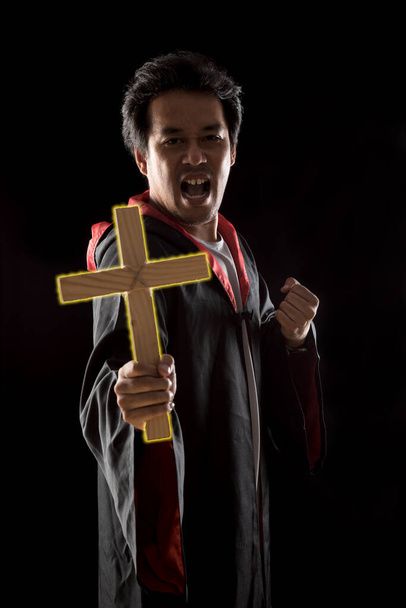 Σκούρο τόνο, φως χείλος - Ιερέας δείχνουν σταυρό φωτισμού, ενώ κρατώντας τη Βίβλο για να εξορκίσει φάντασμα σε μαύρο φόντο - Φωτογραφία, εικόνα