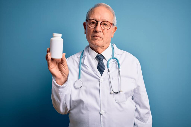 Senior uomo medico dai capelli grigi in possesso di pillole farmaceutiche su sfondo blu con un'espressione fiduciosa sul viso intelligente pensando grave
 - Foto, immagini