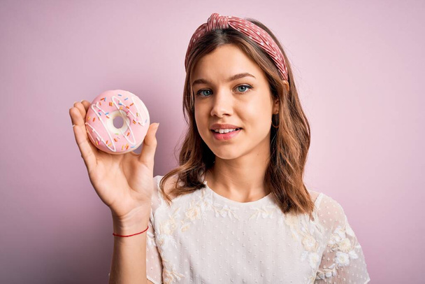 Νεαρό ξανθό κορίτσι τρώει γλυκό ντόνατ ζάχαρη αρτοποιίας πάνω από ροζ απομονωμένο φόντο με ένα χαρούμενο πρόσωπο στέκεται και χαμογελά με αυτοπεποίθηση χαμόγελο δείχνει τα δόντια - Φωτογραφία, εικόνα