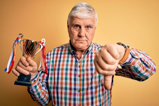 Senior handsome hoary champion man holding trophy award for victory over yellow background with angry face, negative sign showing dislike with thumbs down, rejection concept - Foto, Imagen