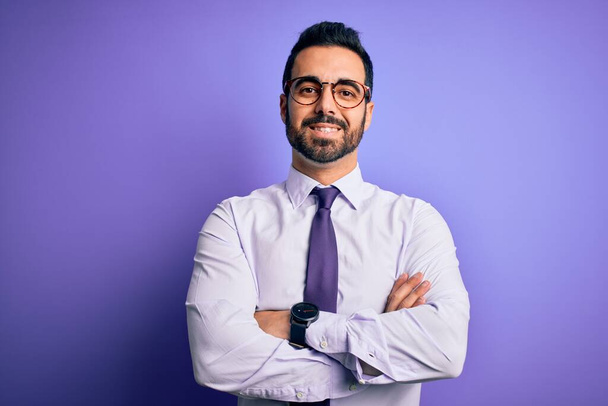 Красивый бизнесмен с бородой в галстуке и очках на фиолетовом фоне счастливое лицо, улыбающееся со скрещенными руками, смотрящее в камеру. Позитивный человек
. - Фото, изображение