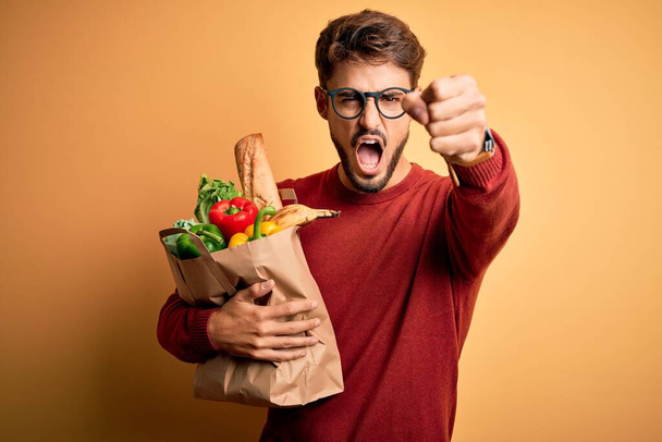 Junger Mann mit Brille, Papiertüte mit Lebensmitteln vor isoliertem gelben Hintergrund, genervt und frustriert schreiend vor Wut, verrückt und schreiend mit erhobener Hand, Wut Konzept - Foto, Bild