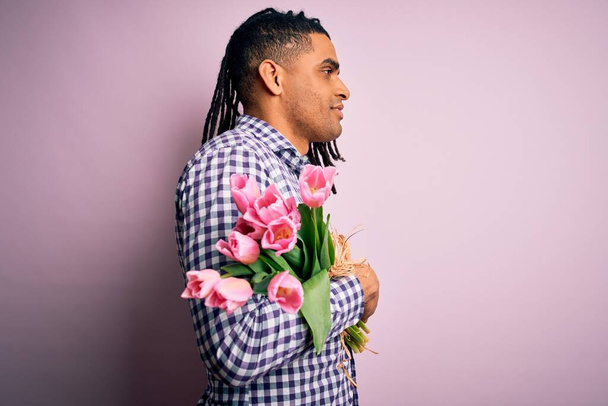 Jonge Afrikaanse amerikaanse afro-romantische man met dreadlocks houden boeket van roze tulpen op zoek naar kant, ontspannen profiel poseren met een natuurlijk gezicht en zelfverzekerde glimlach. - Foto, afbeelding