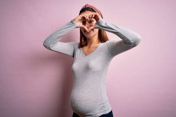 Young beautiful teenager girl pregnant expecting baby over isolated pink background Doing heart shape with hand and fingers smiling looking through sign - Fotografie, Obrázek