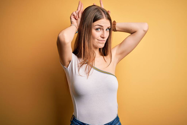Jeune belle femme rousse portant un t-shirt décontracté sur fond jaune isolé Faire des gestes d'oreilles de lapin avec les paumes des mains à l'air cynique et sceptique. Concept lapin de Pâques
. - Photo, image