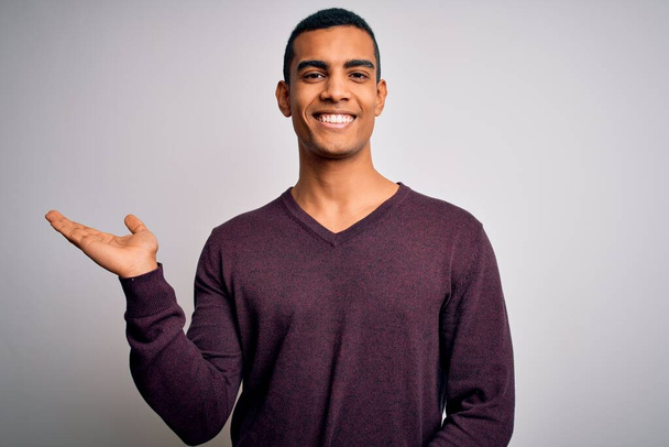 Junger gutaussehender afrikanisch-amerikanischer Mann trägt lässigen Pullover über weißem Hintergrund und lächelt fröhlich und zeigt mit der Handfläche in die Kamera. - Foto, Bild