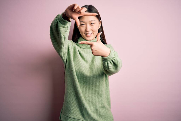 Giovane bella donna asiatica indossa maglione invernale verde su sfondo rosa solated sorridente rendendo cornice con le mani e le dita con il viso felice. Creatività e concetto di fotografia
. - Foto, immagini