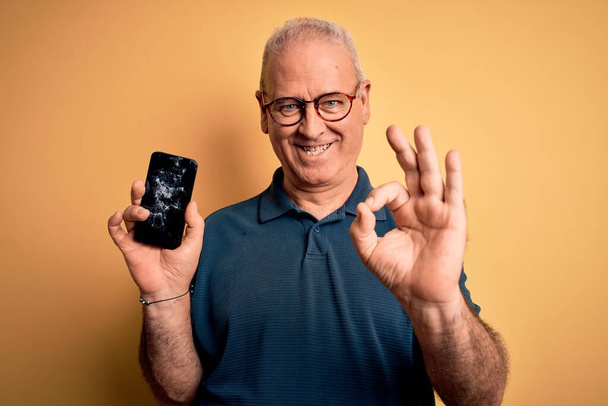 Middle age man holding broken smartphone showing cracked screen over yellow background doing ok sign with fingers, excellent symbol - Fotó, kép