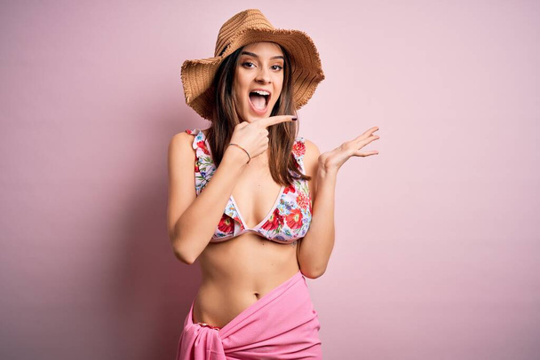 Jonge mooie vrouw op vakantie met bikini en zomerhoed over roze achtergrond verbaasd en glimlachend naar de camera terwijl presenteren met de hand en wijzen met de vinger. - Foto, afbeelding