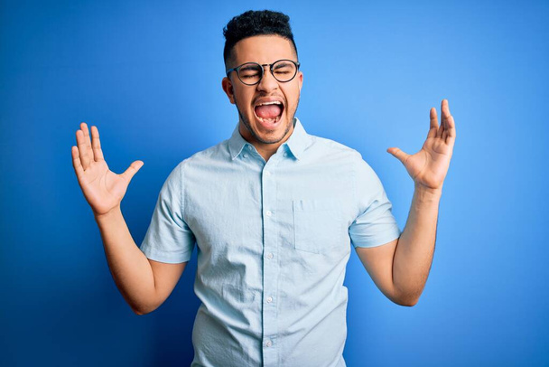 Ein junger gutaussehender Mann in lässigem Sommerhemd und Brille vor isoliertem blauen Hintergrund feiert verrückt und erfolgsverrückt mit erhobenen Armen und geschlossenen Augen, die aufgeregt schreien. Siegerkonzept - Foto, Bild