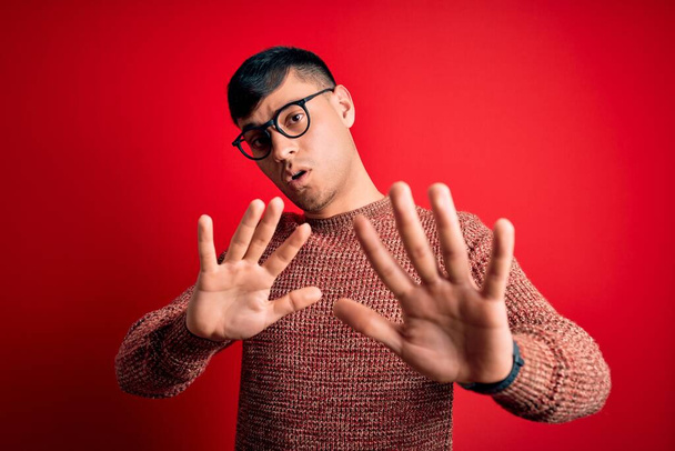 Homem hispânico bonito jovem usando óculos nerd sobre fundo vermelho Movendo as mãos longe palmas mostrando recusa e negação com expressão assustadora e nojenta. Pare e proibido
. - Foto, Imagem