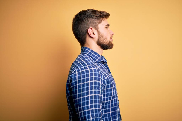 Young blond businessman with beard and blue eyes wearing shirt over yellow background looking to side, relax profile pose with natural face and confident smile. - 写真・画像