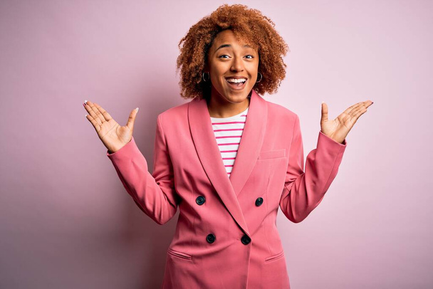Joven hermosa mujer de negocios afroamericana con el pelo rizado usando elegante chaqueta rosa celebrando loco y sorprendido por el éxito con los brazos levantados y los ojos abiertos gritando emocionados. Concepto ganador
 - Foto, Imagen