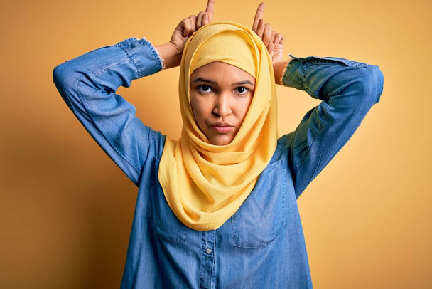 Mladá krásná žena s kudrnaté vlasy nosí arabsky tradiční hidžáb přes žluté pozadí dělá legrační gesto s prstem přes hlavu jako býčí rohy - Fotografie, Obrázek