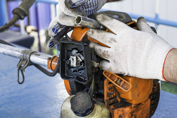 мастер в рабочих перчатках ремонтирует двигатель старого газового триммера и отвинчивает свечу зажигания гаечным ключом
 - Фото, изображение