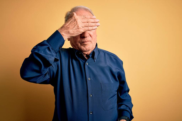 Grey haired senior man wearing casual blue shirt standing over yellow background covering eyes with hand, looking serious and sad. Sightless, hiding and rejection concept - Fotografie, Obrázek