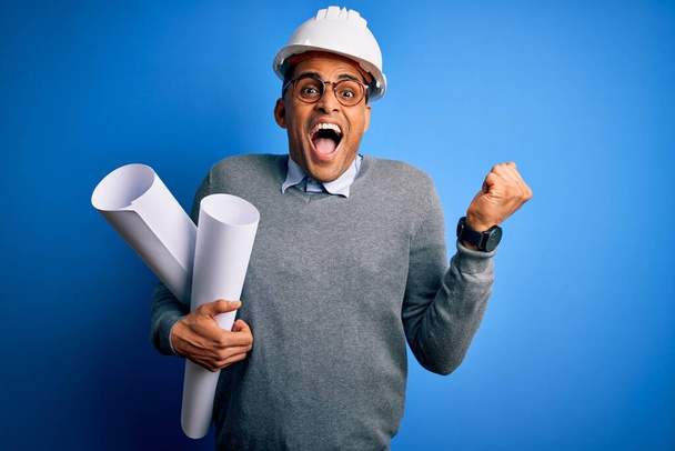 Молодий африканський архітектор, одягнений в шолом безпеки, з синіми малюнками, що кричать гордо і відзначають перемогу і успіх дуже схвильовані, підбадьорюючи емоції. - Фото, зображення