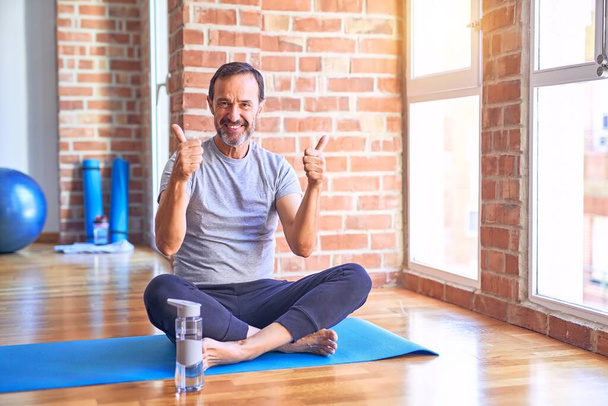 Średniego wieku przystojny sportowiec siedzi na macie robi rozciąganie ćwiczenia jogi na siłowni znak sukcesu robi pozytywny gest z ręki, kciuki do góry uśmiechnięty i szczęśliwy. Radosna ekspresja i gest zwycięzcy. - Zdjęcie, obraz