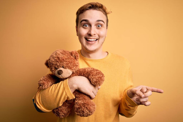 Jeune homme rousse beau tenant ours en peluche jouet animal sur fond jaune Sourire heureux pointant avec la main et le doigt sur le côté
 - Photo, image
