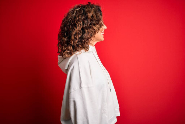 Średniego wieku piękna sportowa kobieta w białej bluzie na izolowanym czerwonym tle patrząc na bok, relaks profil pozować z naturalną twarz z pewnym uśmiechem. - Zdjęcie, obraz