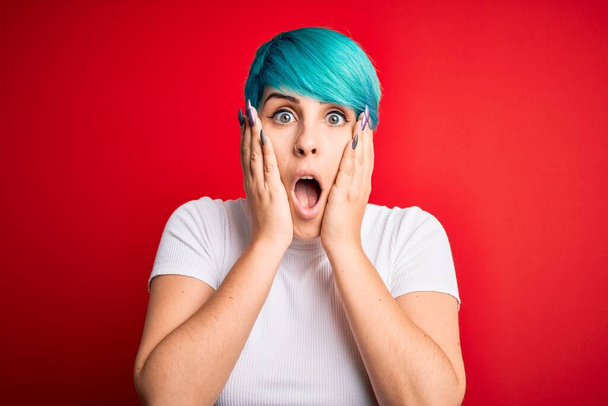 Νεαρή όμορφη γυναίκα με μπλε μαλλιά μόδας φορώντας casual t-shirt πάνω από το κόκκινο φόντο φοβισμένος και σοκαρισμένος, έκπληξη και κατάπληκτος έκφραση με τα χέρια στο πρόσωπο - Φωτογραφία, εικόνα