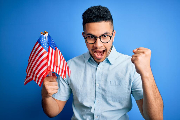 Jonge knappe patriottische man die Amerikaanse vlaggen vasthoudt... die de onafhankelijkheidsdag viert... trots schreeuwend en de overwinning en het succes viert... erg opgewonden, juichende emotie. - Foto, afbeelding
