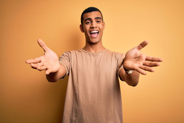 Jonge knappe Afro-Amerikaanse man met casual t-shirt over gele achtergrond kijkend naar de camera glimlachend met open armen voor knuffel. Vrolijke uitdrukking die geluk omarmt. - Foto, afbeelding