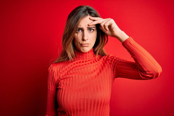 Junge schöne brünette Frau trägt lässigen Rollkragenpullover über rotem Hintergrund besorgt und gestresst über ein Problem mit der Hand auf der Stirn, nervös und krisenängstlich - Foto, Bild