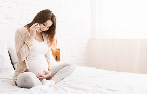 Έγκυος γυναίκα μιλάει στο τηλέφωνο και αγκαλιάζει την κοιλιά της στο σπίτι - Φωτογραφία, εικόνα