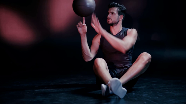 Αθλητής με σταυρωμένα πόδια γυρίζοντας μπάλα και κοιτάζοντας κάμερα στο σκοτάδι - Πλάνα, βίντεο