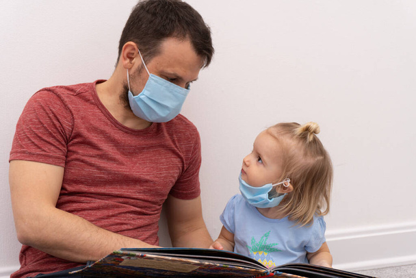 Girl in medical mask looks at father who is reading book - Foto, Imagen