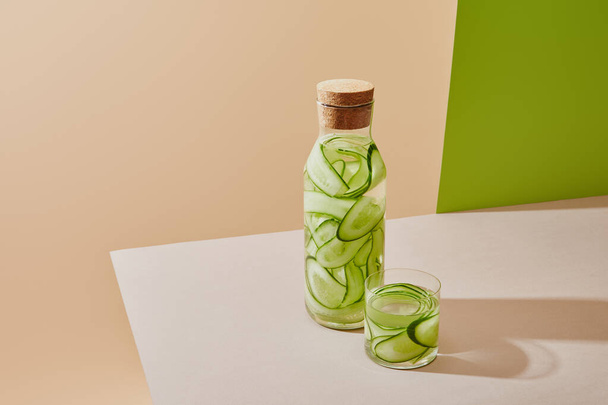 high angle view of glass and bottle with cork filled with fresh water and sliced cucumbers on table on beige and green background - Photo, image