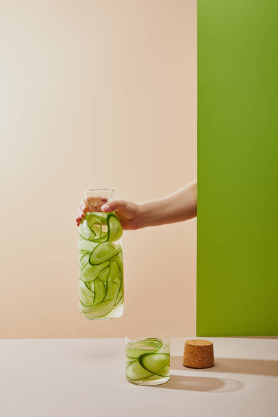 Cropped view of female hand holding bottle filled with water and sliced cucumbers on beige and green background - Fotó, kép