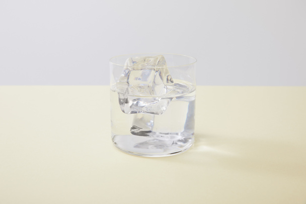 Glass with water and ice cubes on yellow surface isolated on grey - 写真・画像