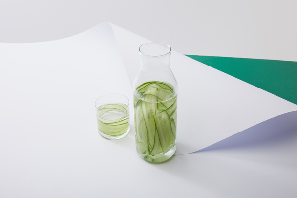 nagy látószögű kilátás palack és üveg vízzel és szeletelt uborka fehér papír felületen szürke és zöld háttér  - Fotó, kép