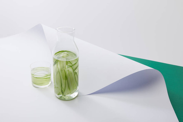 high angle view of bottle and glass with fresh drink and sliced cucumbers on white paper surface on grey and green background  - Foto, immagini