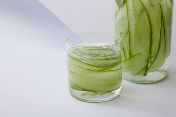 Close up view of bottle and glass with fresh drink and sliced cucumbers on grey and white background - Foto, Imagen