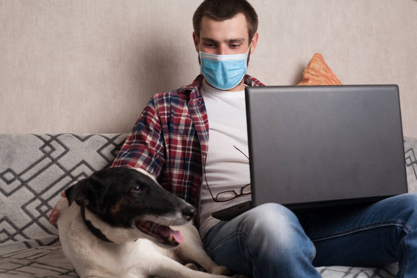 Adam evcil hayvanıyla birlikte evde çalışıyor. Adam tıbbi maskeli bir köpekle kanepede iletişim kuruyor.. - Fotoğraf, Görsel