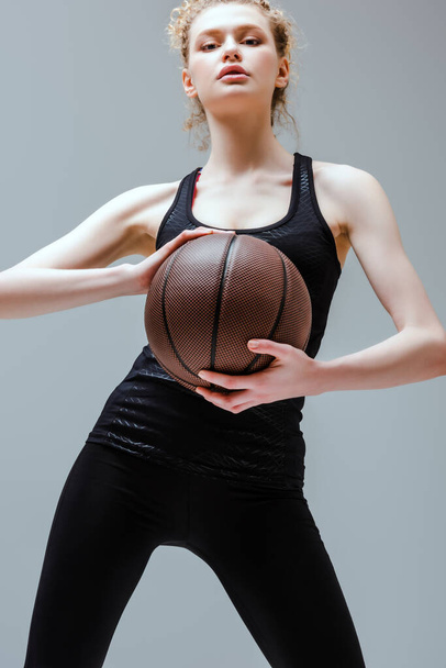 curly woman in sportswear holding basketball isolated on grey - Zdjęcie, obraz