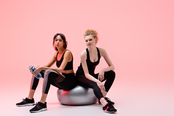 sportive girl sitting on fitness mall with african american woman holding sports bottle on pink  - Fotoğraf, Görsel