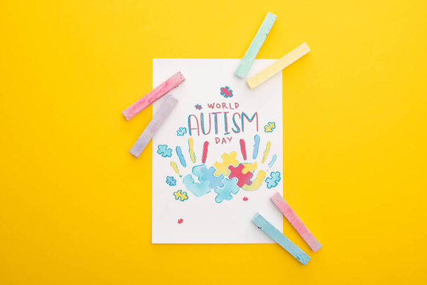 top view of card with puzzle and hand prints for World Autism Day isolated on yellow with chalks - Fotografie, Obrázek