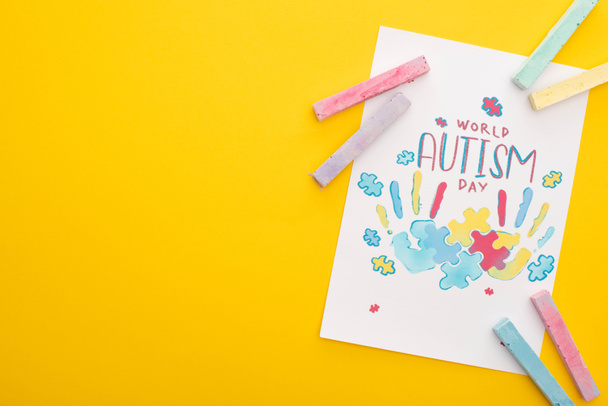 top view of card with puzzle and hand prints for World Autism Day isolated on yellow with colorful chalks - Foto, afbeelding